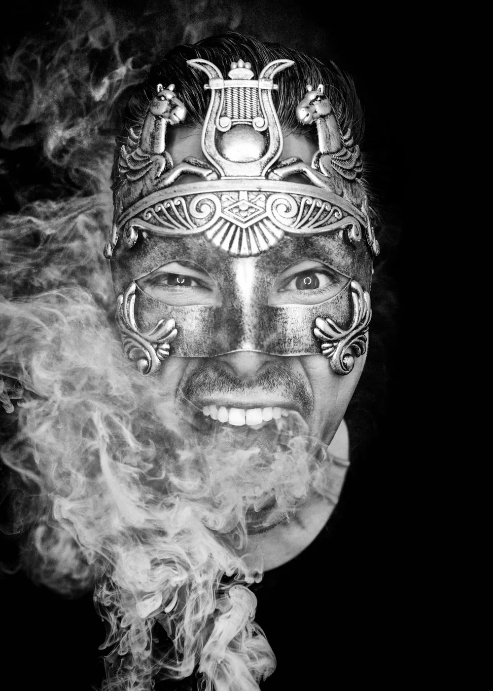 A person wearing an ornate, metallic mask with decorative details and two animal figures on the forehead, smiling with teeth showing, surrounded by smoke.
