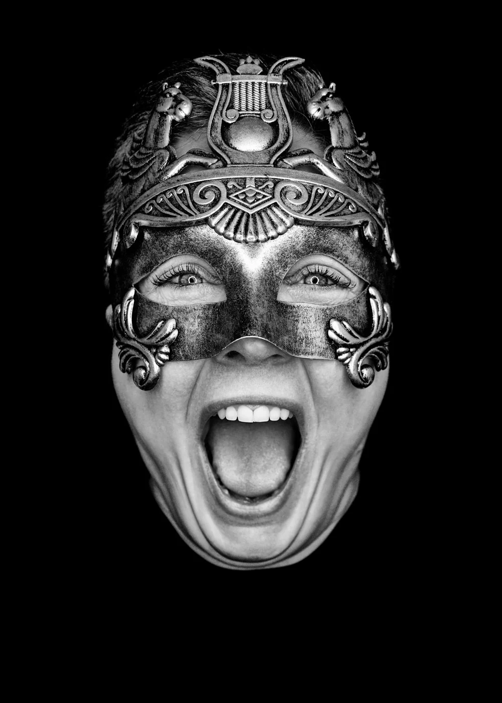Black and white photo of a person wearing an ornate metallic mask with a lion and lyre design, showing a surprised or shouting expression.