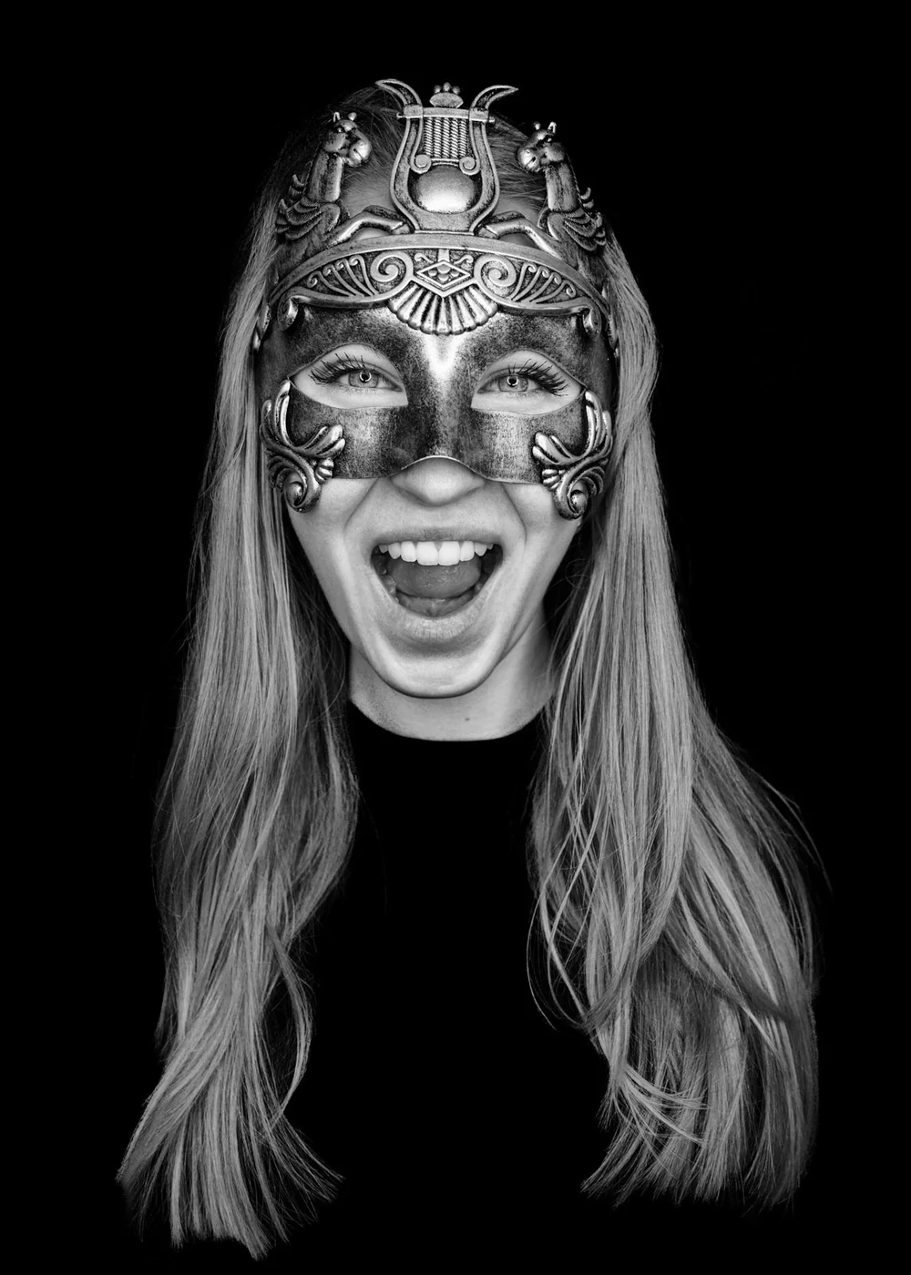 A woman with long hair wearing a decorative mask with intricate designs and open mouth, smiling against a black background.