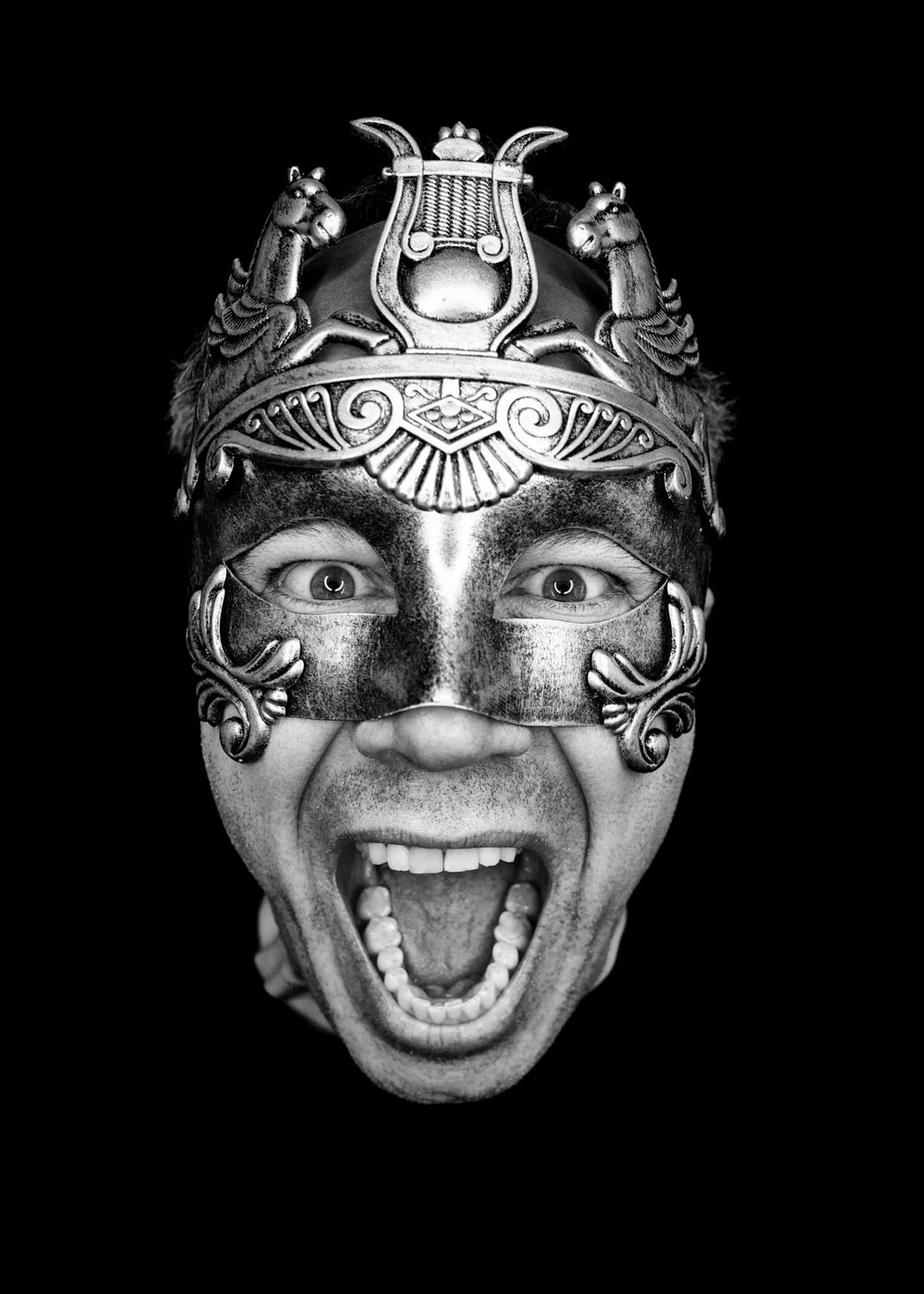 Person wearing a decorative mask with a humorous facial expression, mouth open in a yell or laugh, against a black background.
