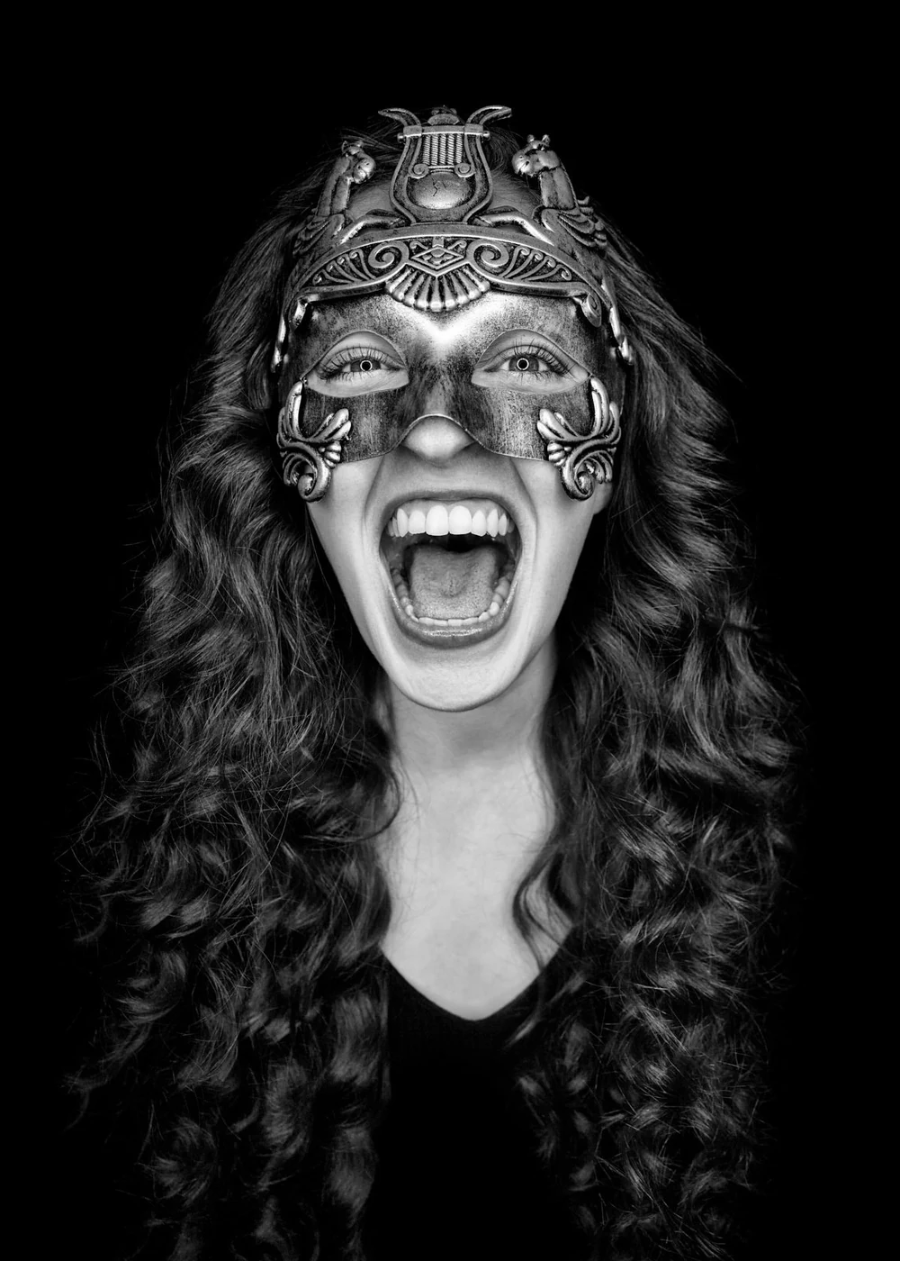 A woman with long curly hair wearing an ornate mask, shouting with her mouth open wide, in black and white.