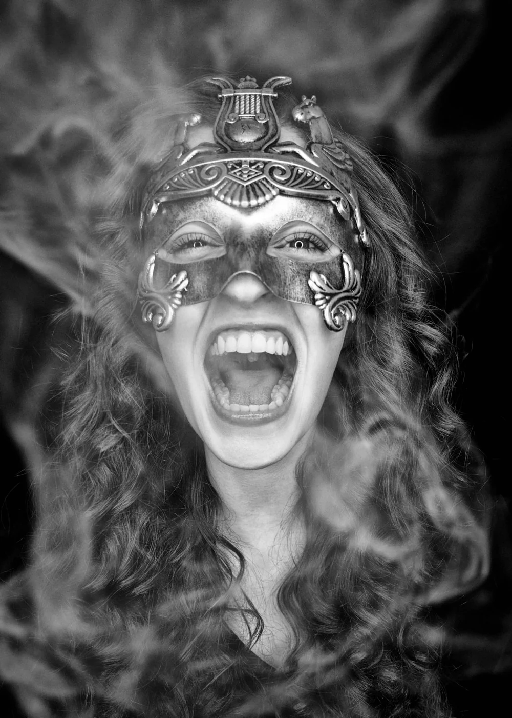 A woman with curly hair wearing an ornate masquerade mask, screaming with her mouth wide open, in black and white.
