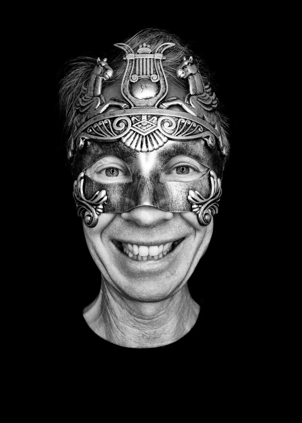 Person smiling wearing an ornate mask with classical and mythical designs against a black background.