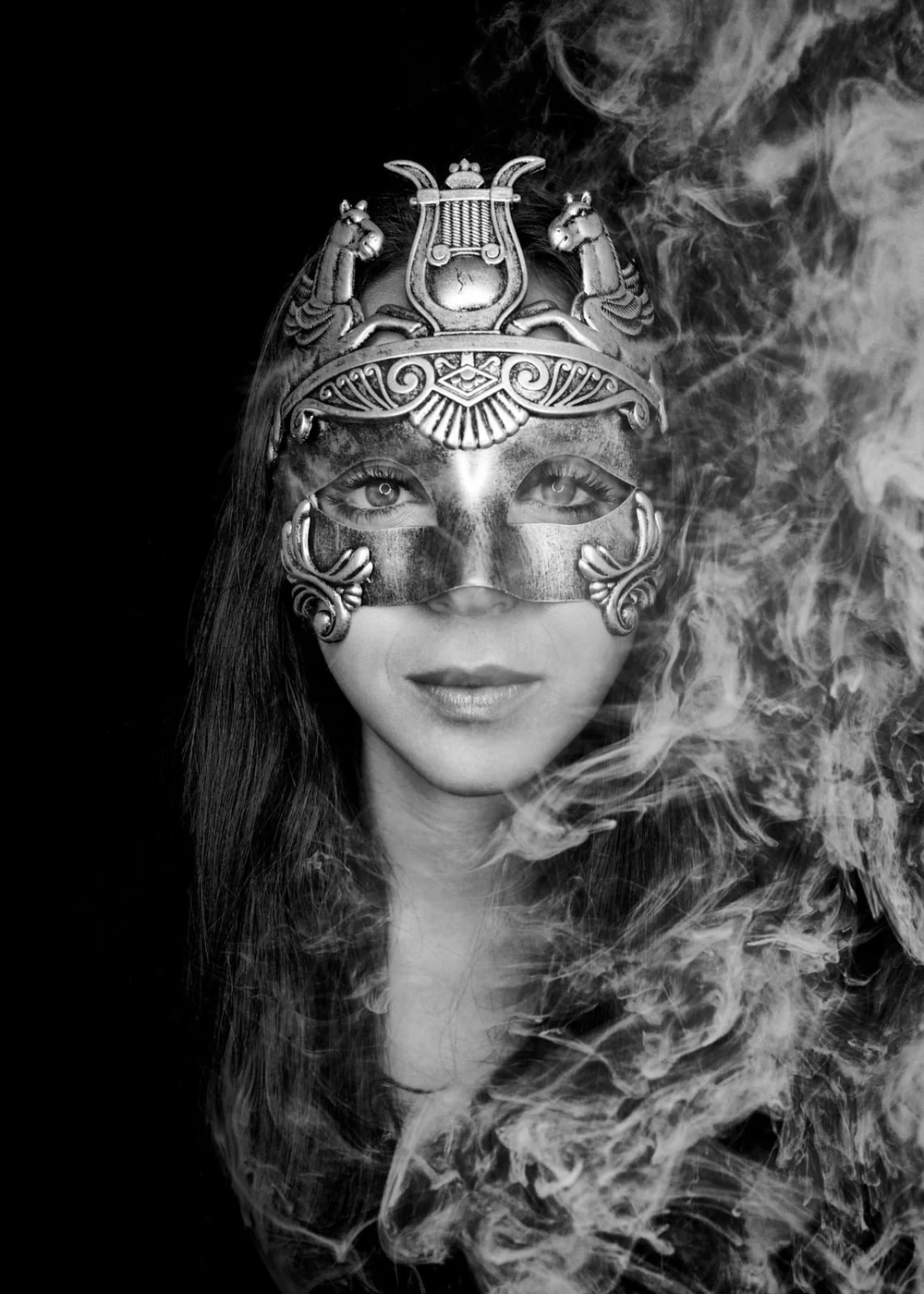 A woman wearing an ornate, metallic mask with intricate designs and animal figures, surrounded by swirling smoke against a dark background.