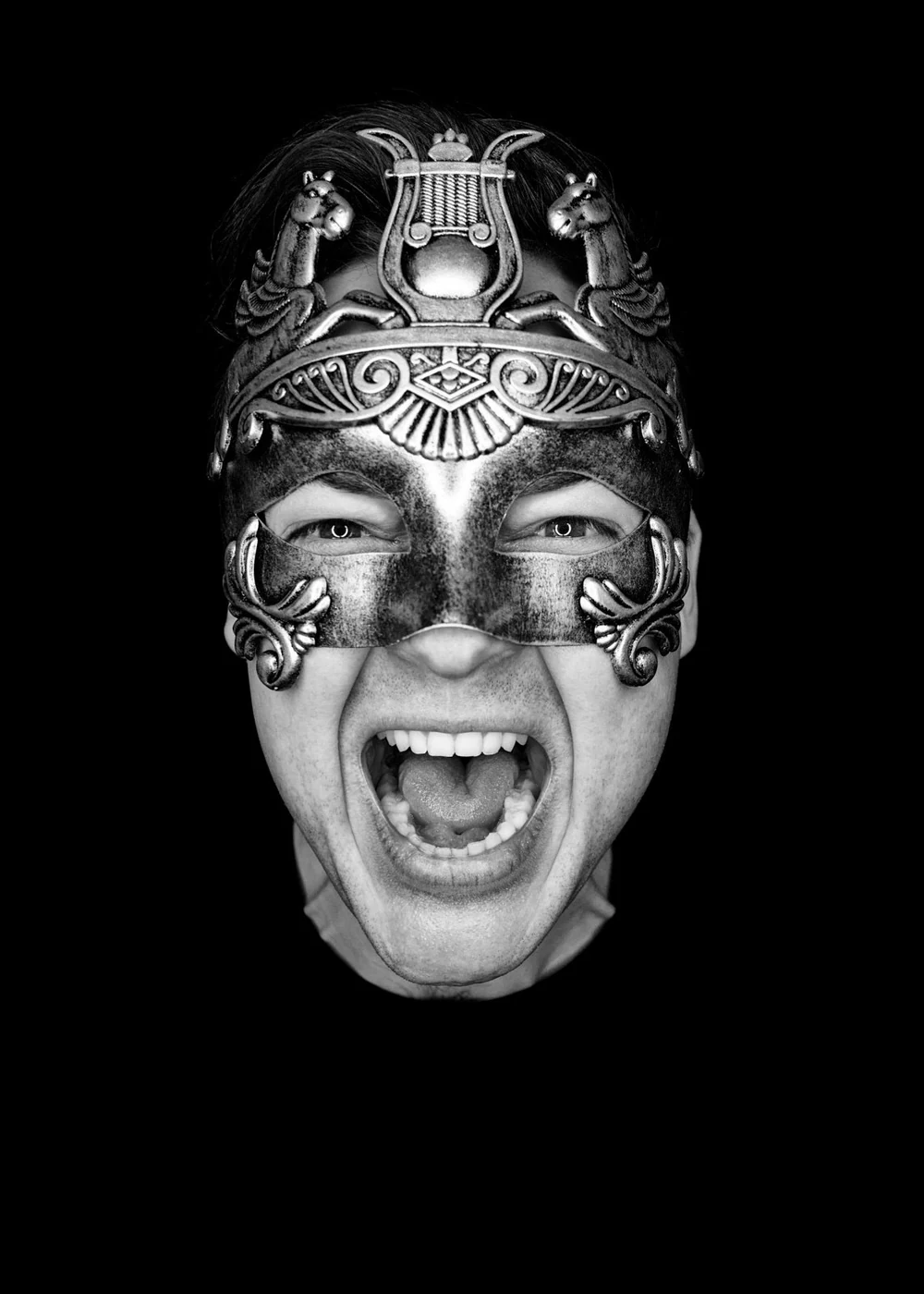 A person wearing an ornate metal mask with animal and mythical creature designs, shouting with mouth open and tongue out, against a black background.