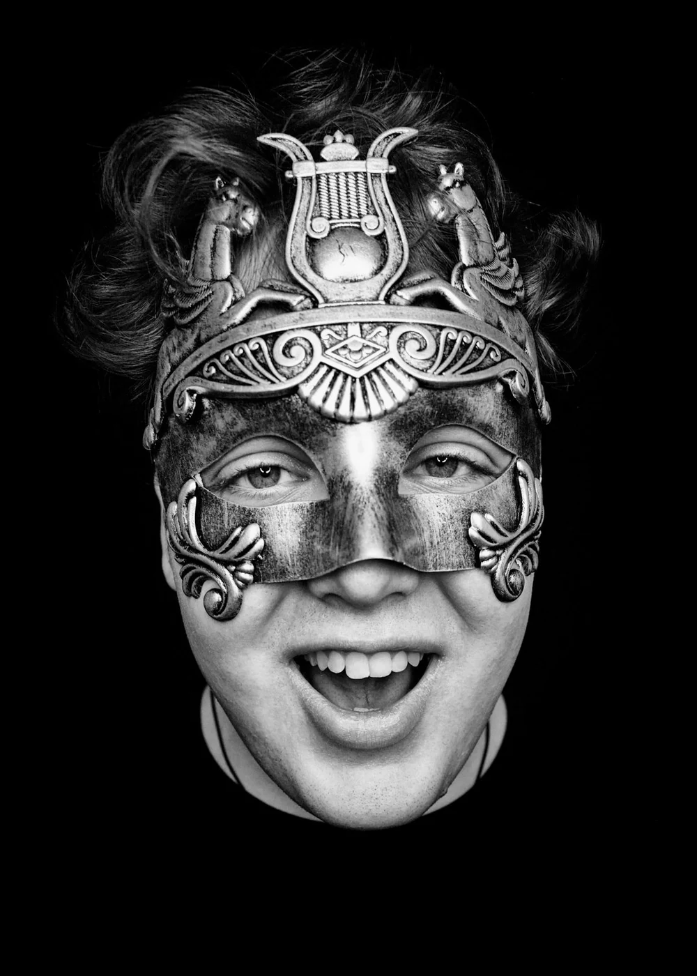 A person wearing an ornate metal mask with intricate designs and waves, covering the upper half of their face, smiling with an open mouth, black background.