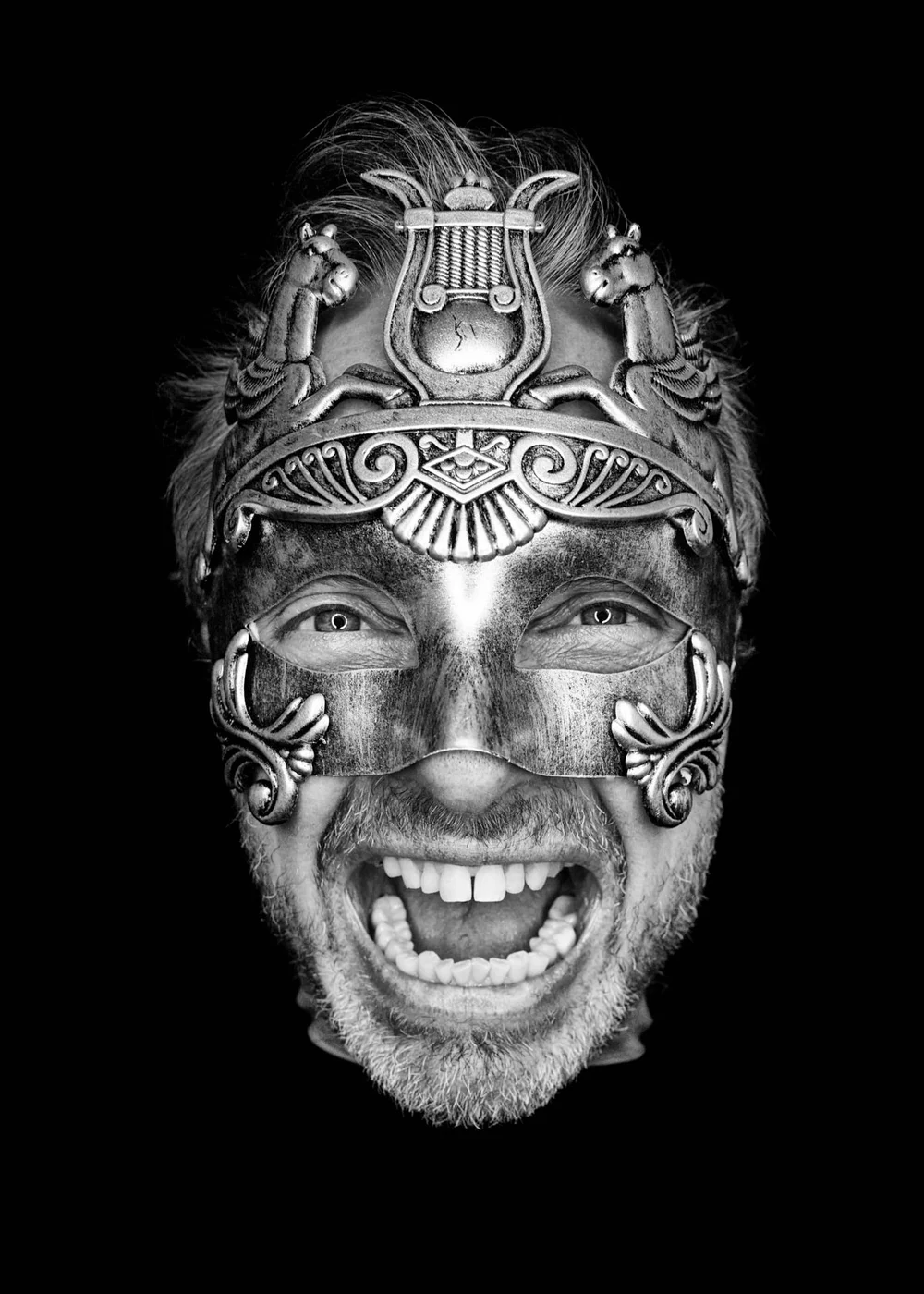 Black and white photo of a man with gray hair and beard wearing an ornate metallic mask and headdresses, smiling widely with eyes visible through the mask.
