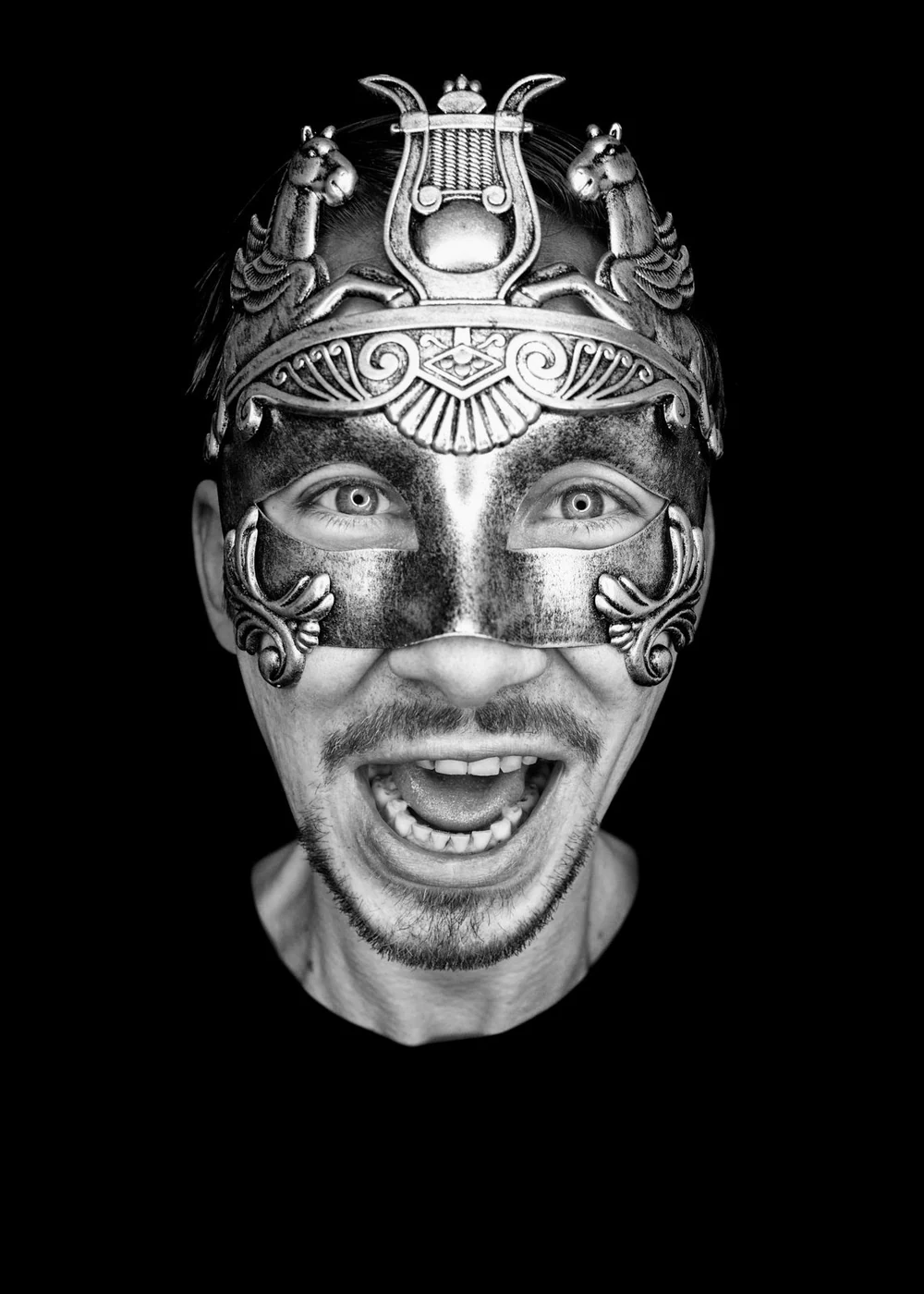 Black and white photo of a man wearing an ornate, decorative mask with lion and horse figures, smiling and showing his teeth, against a black background.