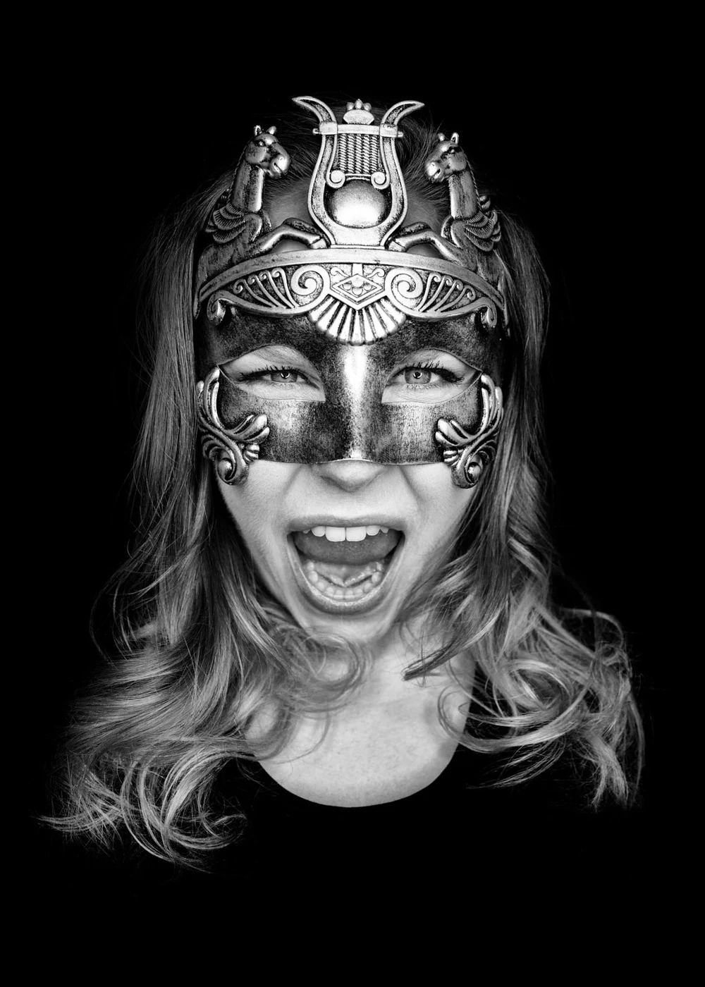 A woman with wavy hair wearing a decorative mask covering her eyes and nose, with her mouth open in a joyful expression, against a black background.