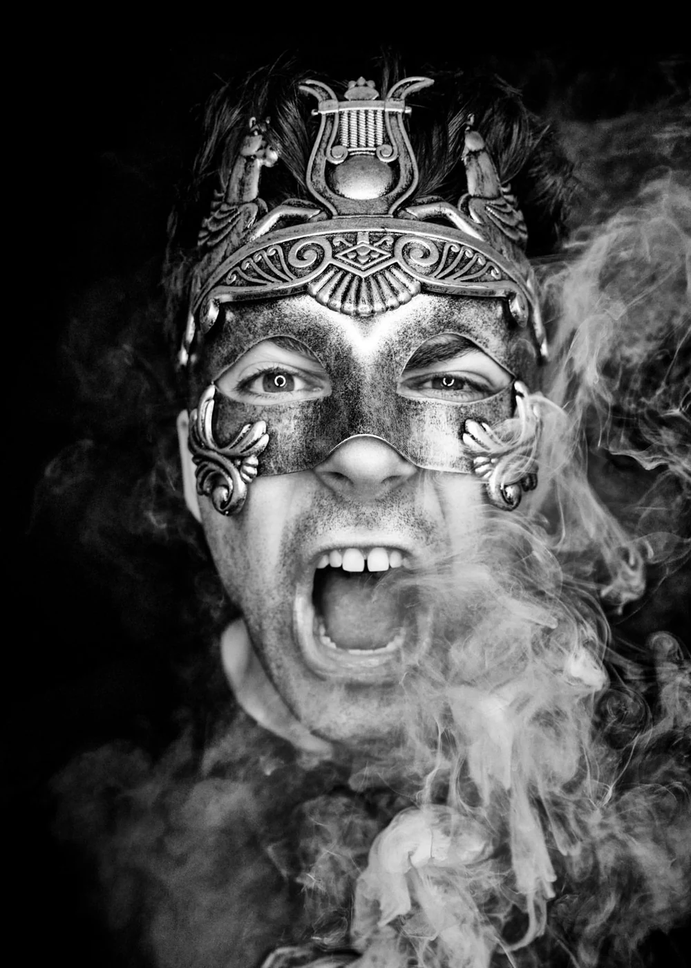 Person wearing an ornate mask with a headdress, shouting with mouth open, surrounded by smoke in black and white.