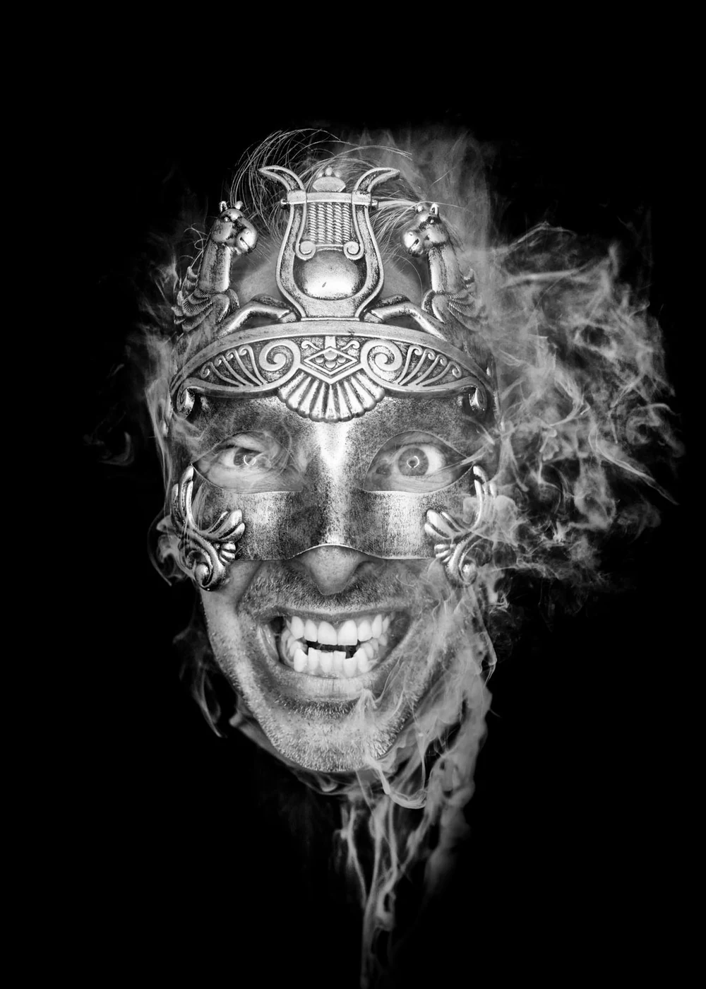 A person smiling with visible teeth wearing a decorative metal mask and an ornate headdress, surrounded by swirling smoke against a black background.