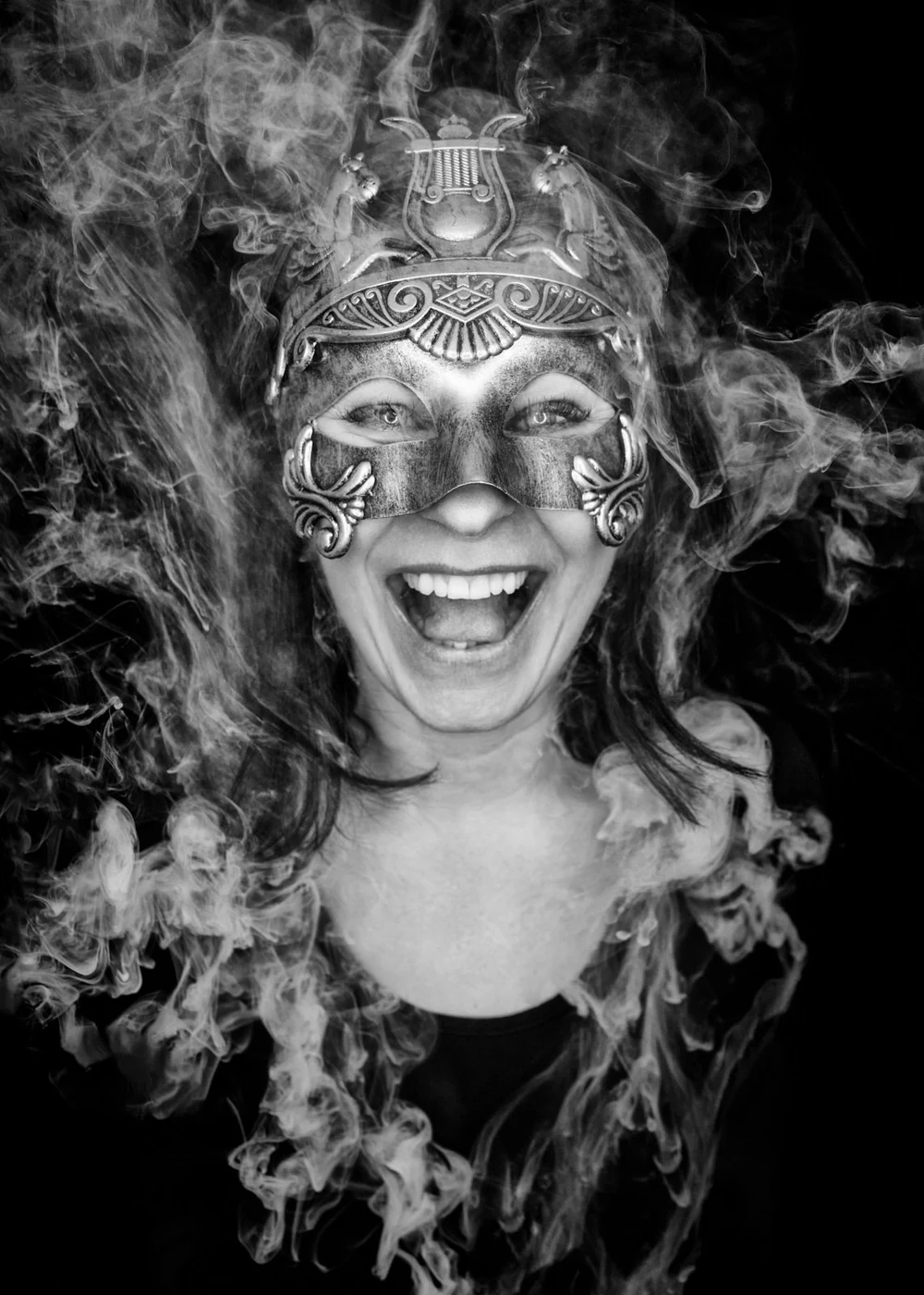 A woman wearing an ornate mask with a ridged, metallic headpiece and wide smile, surrounded by swirling smoke or fog, in black and white.