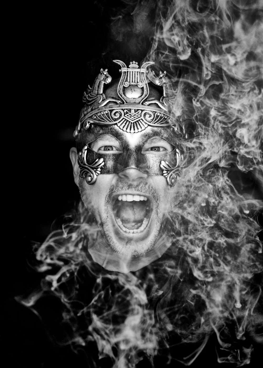 A man wearing a decorative mask with intricate designs and animal figures, shouting with an open mouth, surrounded by swirling smoke or fog against a black background.